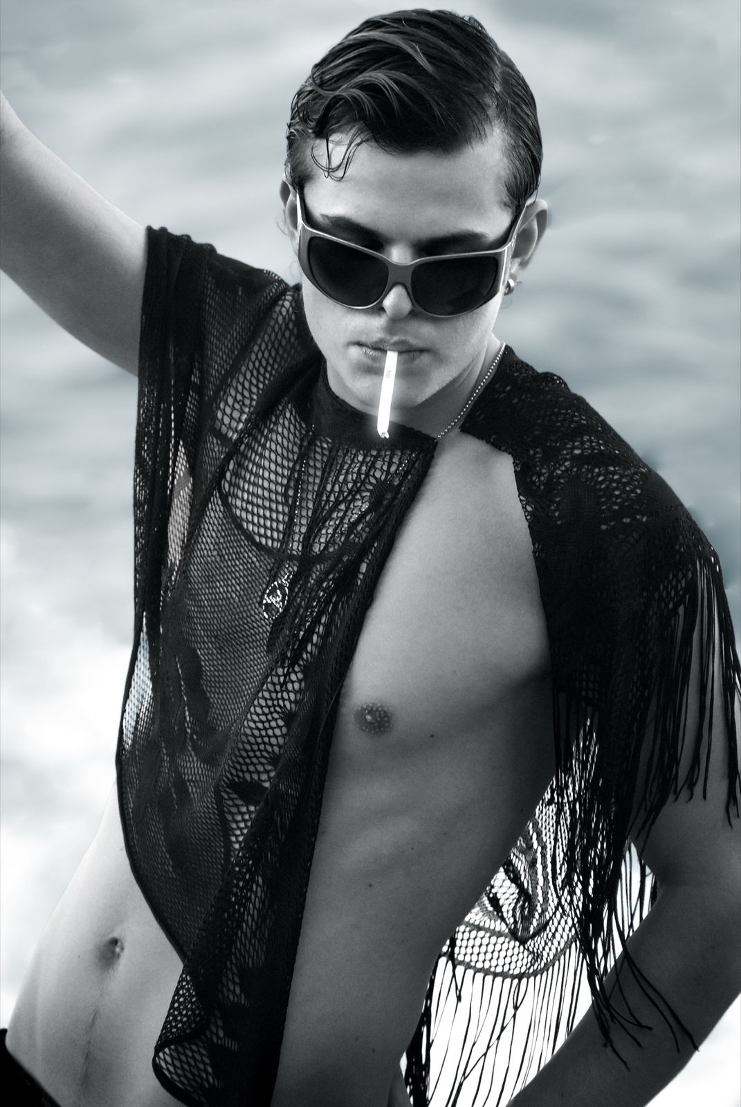 Black and white portrait from a JIGGY shoot: a model in a fringed mesh top and sunglasses, smoking, shot on the water's edge